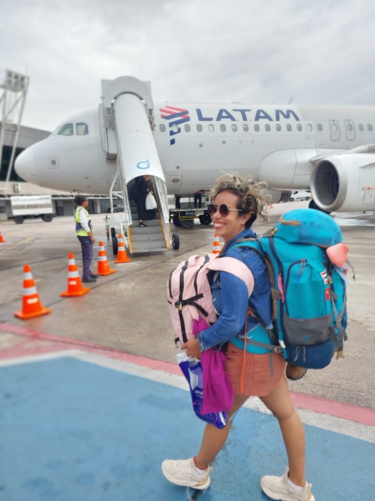 Nanda Alcantara com as mochilas em frente a um aviao representando "a mulher guerreira" carregando muitas coisas na mochila.